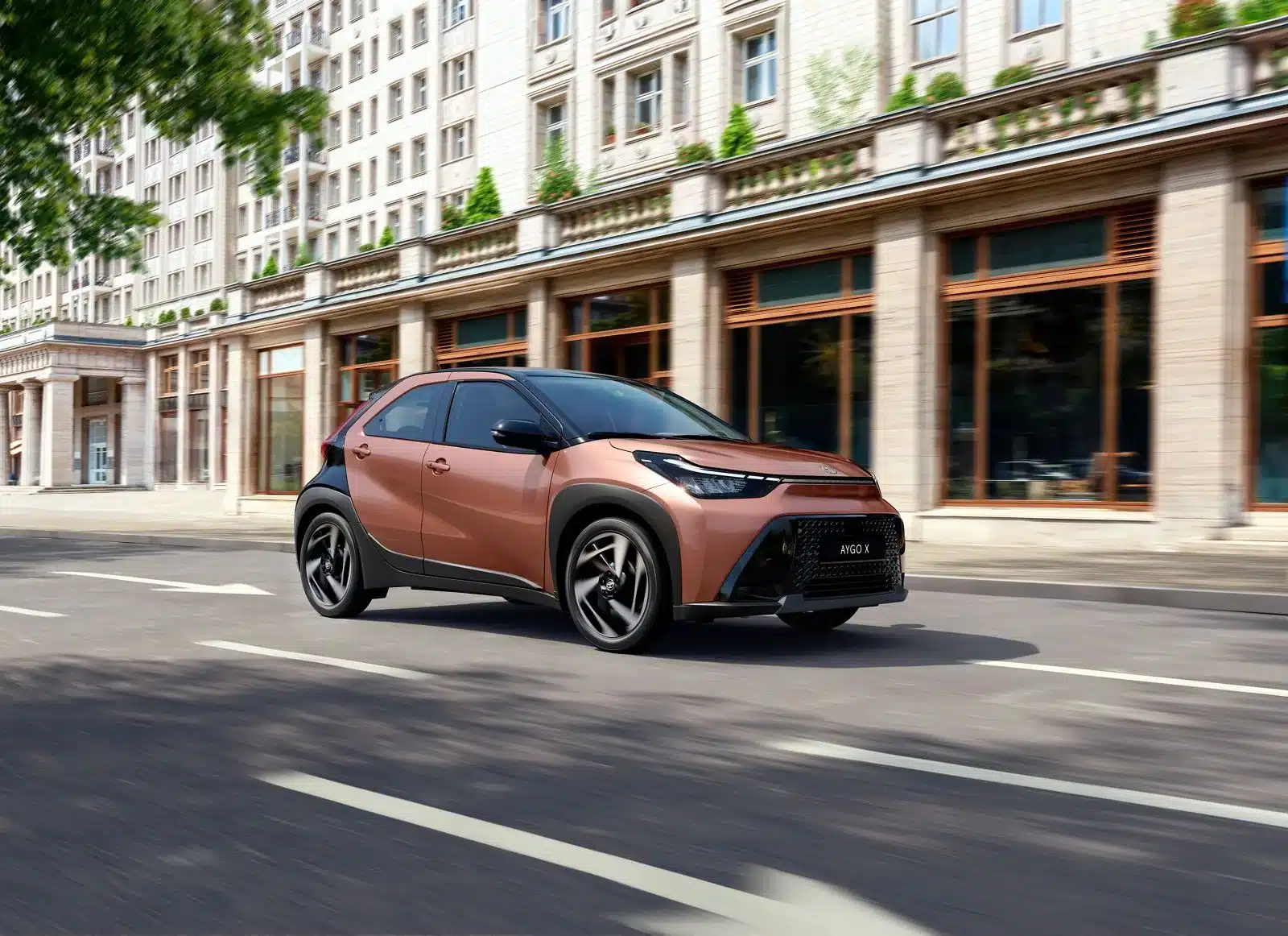 Toyota Aygo X Hybrid fährt dynamisch auf einer breiten Straße in der Stadt.