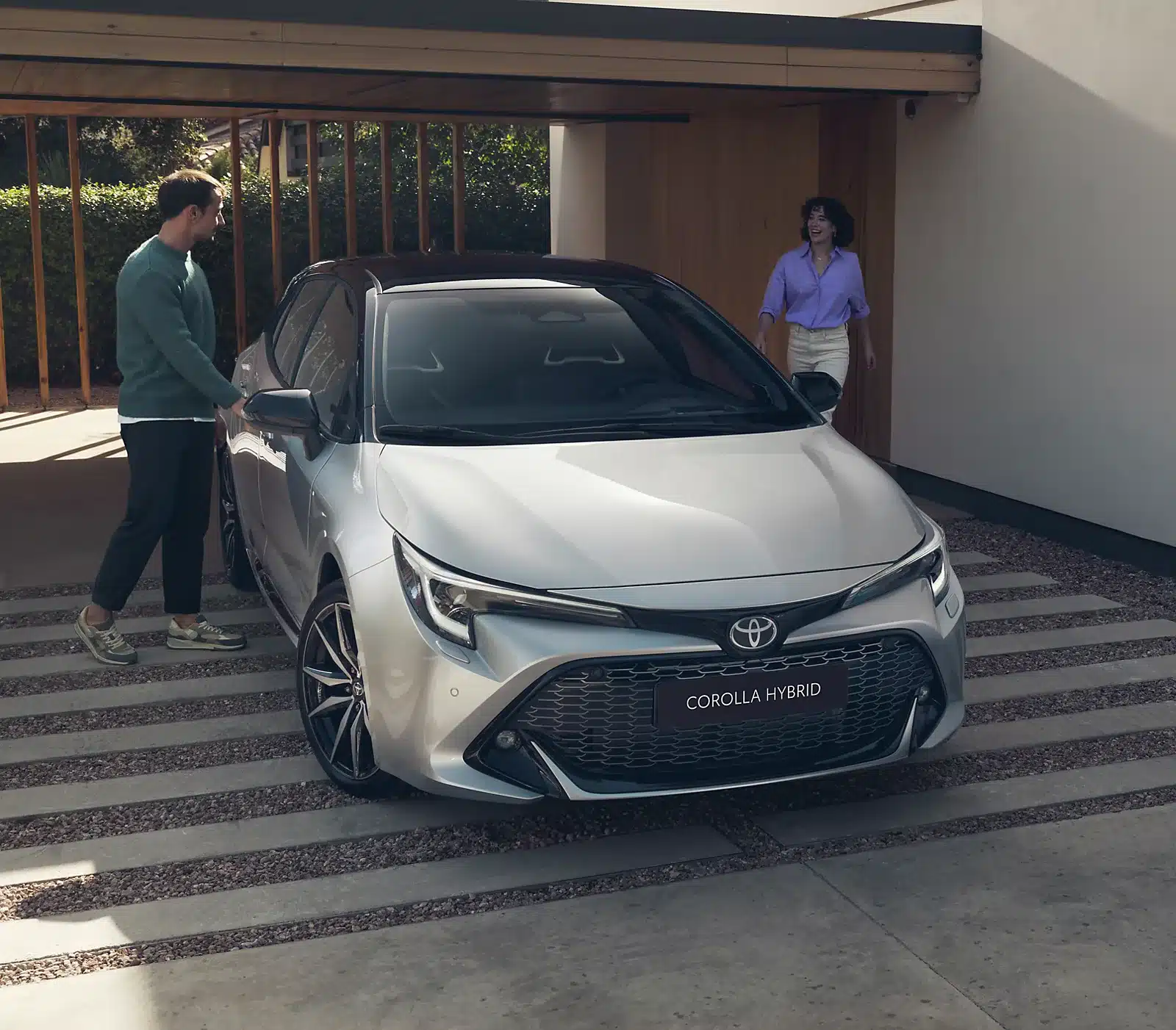 Der Toyota Corolla Hybrid in Silber steht vor einer modernen Garage, ein Mann steht an der Fahrertür, eine Frau kommt lachend hinzu.