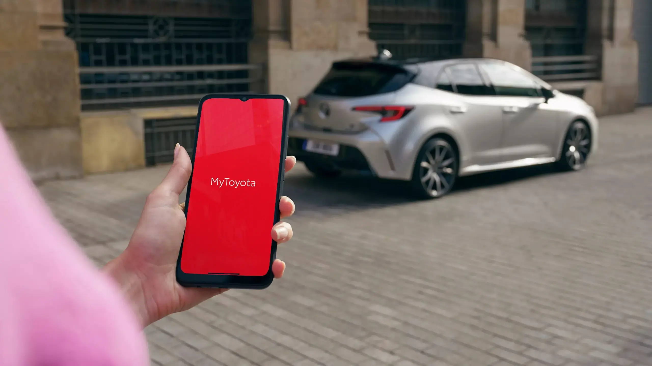 Smartphone mit geöffneter MyToyota App vor einem geparkten silbernen Toyota Corolla Hybrid