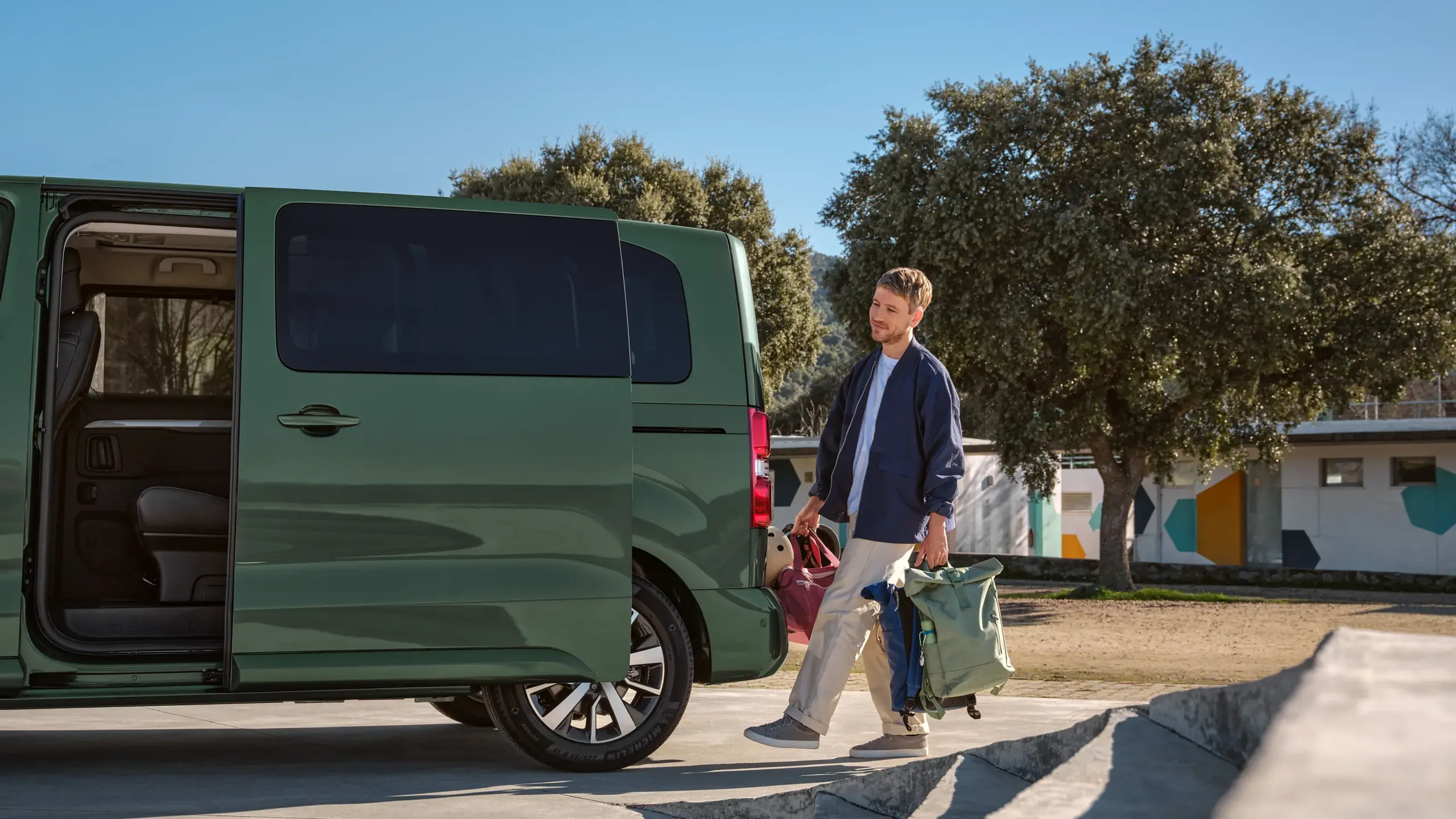 Ein Mann verstaut Taschen im Kofferraum eines grünen Toyota Proace Verso Vans, auf einem sonnigen Parkplatz mit Bäumen.