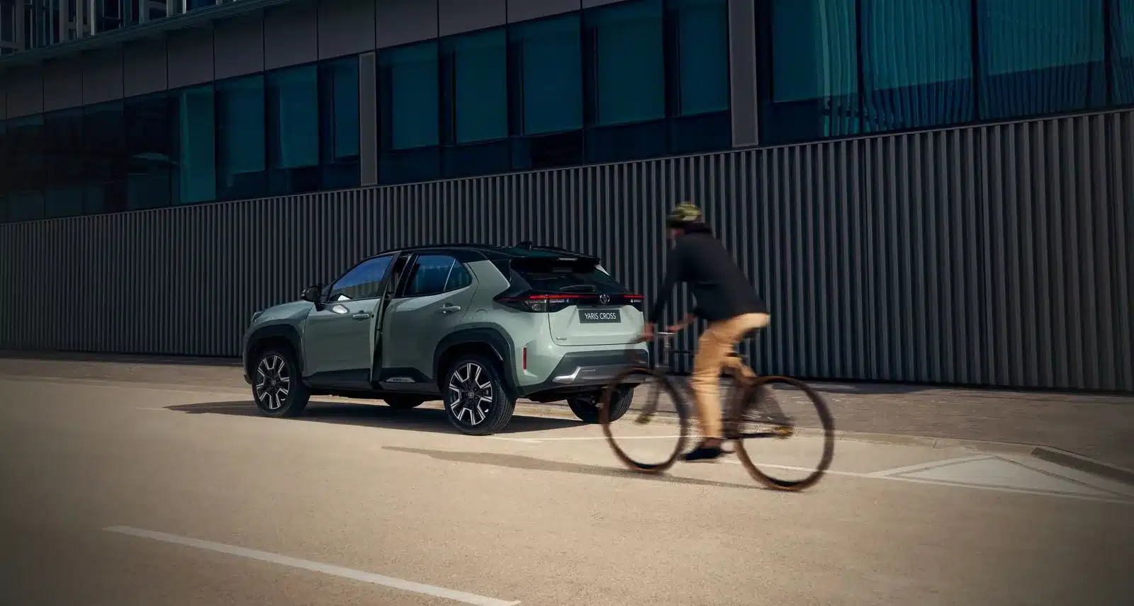 Toyota Yaris Cross als Kompakt-SUV in der Stadt, Blick auf die Fahrzeugseite und das Heck, geöffnete Tür, moderner Hintergrund, Fahrradfahrer fährt vorbei