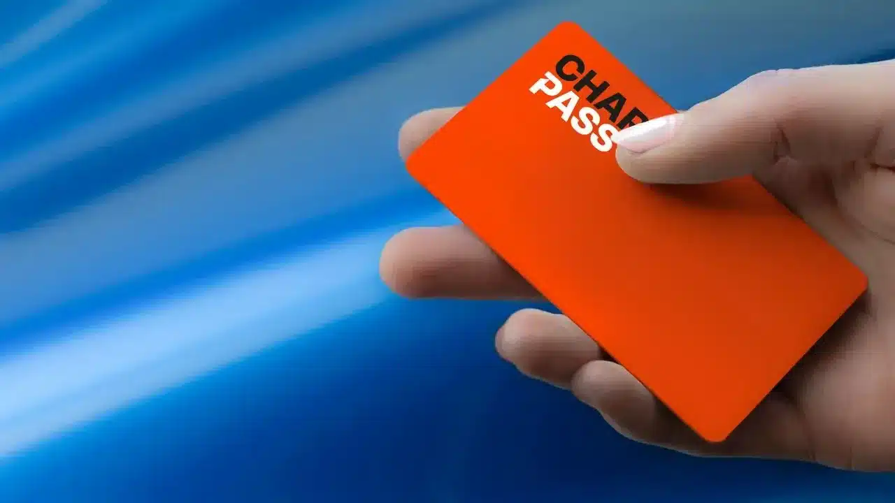 Hand hält eine orangefarbene Ladekarte mit der Aufschrift 'CHARGEPASS' vor verschwommenem blauem Hintergrund.