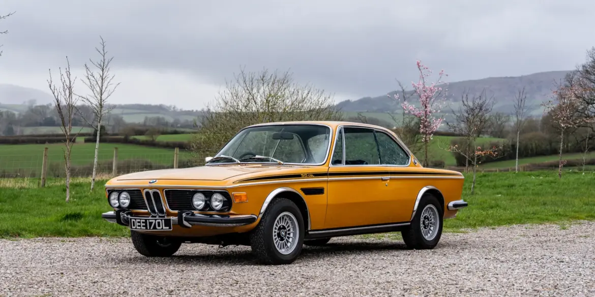 BMW 3.0 CSL Baujahr 1973 in Gelb, Seitenansicht auf Kiesweg, Hintergrund grüne Landschaft