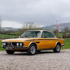 BMW 3.0 CSL Baujahr 1973 in Gelb, Seitenansicht auf Kiesweg, Hintergrund grüne Landschaft