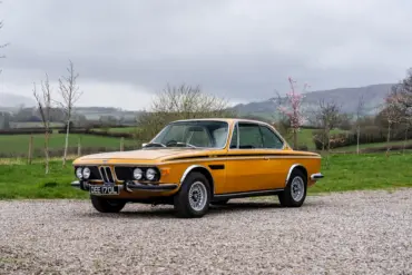 BMW 3.0 CSL Baujahr 1973 in Gelb, Seitenansicht auf Kiesweg, Hintergrund grüne Landschaft