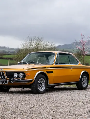 BMW 3.0 CSL Baujahr 1973 in Gelb, Seitenansicht auf Kiesweg, Hintergrund grüne Landschaft
