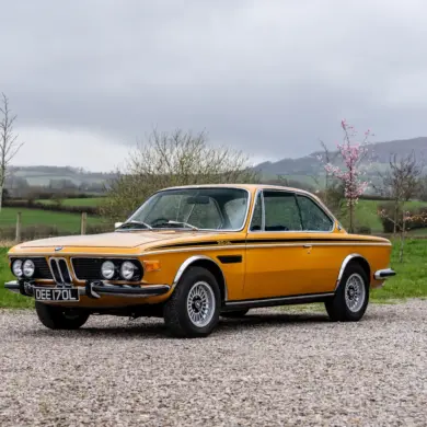 BMW 3.0 CSL Baujahr 1973 in Gelb, Seitenansicht auf Kiesweg, Hintergrund grüne Landschaft