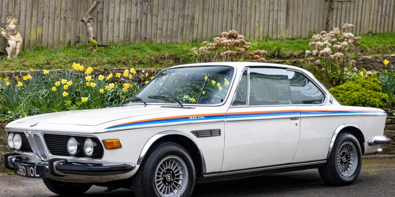Weißer BMW 3.0 CSL von 1973 als rechtsgelenktes Coupé, geparkt vor einem Holzzaun mit Blumen.
