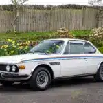Weißer BMW 3.0 CSL von 1973 als rechtsgelenktes Coupé, geparkt vor einem Holzzaun mit Blumen.