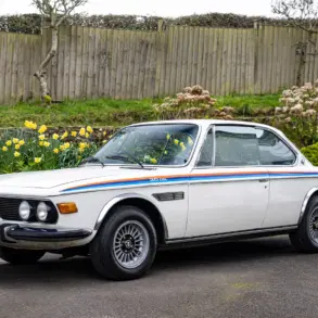 Weißer BMW 3.0 CSL von 1973 als rechtsgelenktes Coupé, geparkt vor einem Holzzaun mit Blumen.
