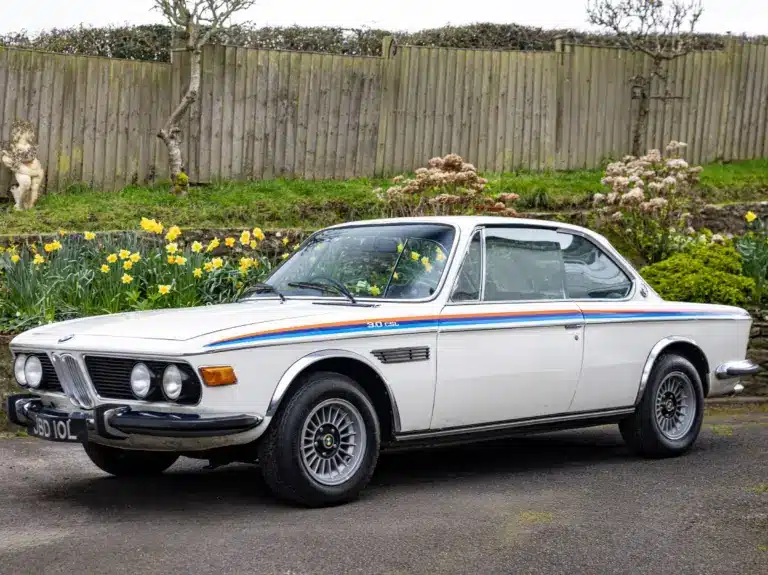 Weißer BMW 3.0 CSL von 1973 als rechtsgelenktes Coupé, geparkt vor einem Holzzaun mit Blumen.