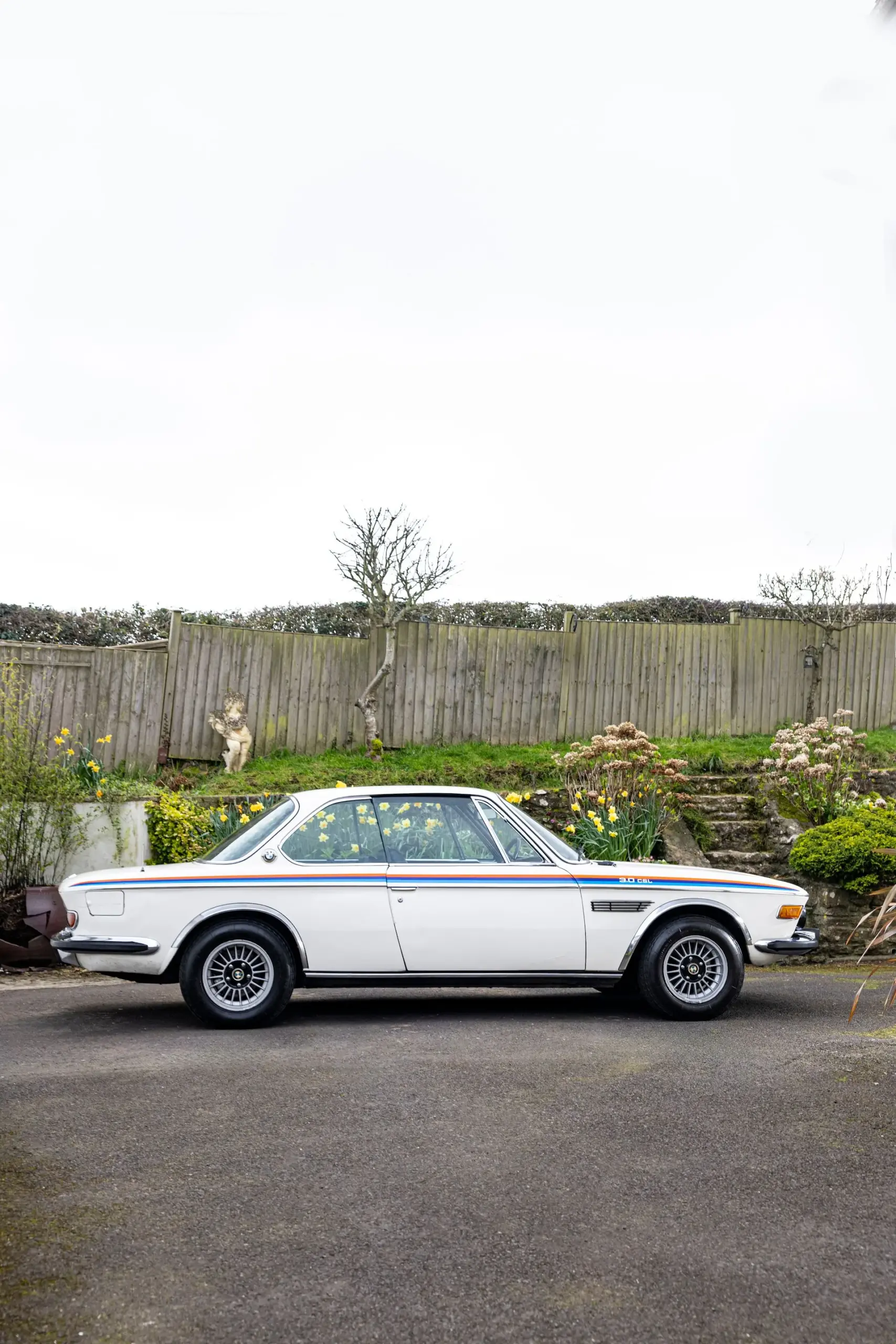 BMW 3.0 CSL, Baujahr 1973, weiß, Seitenansicht, mit M-Streifen, geparkt vor einem Gartenzaun.