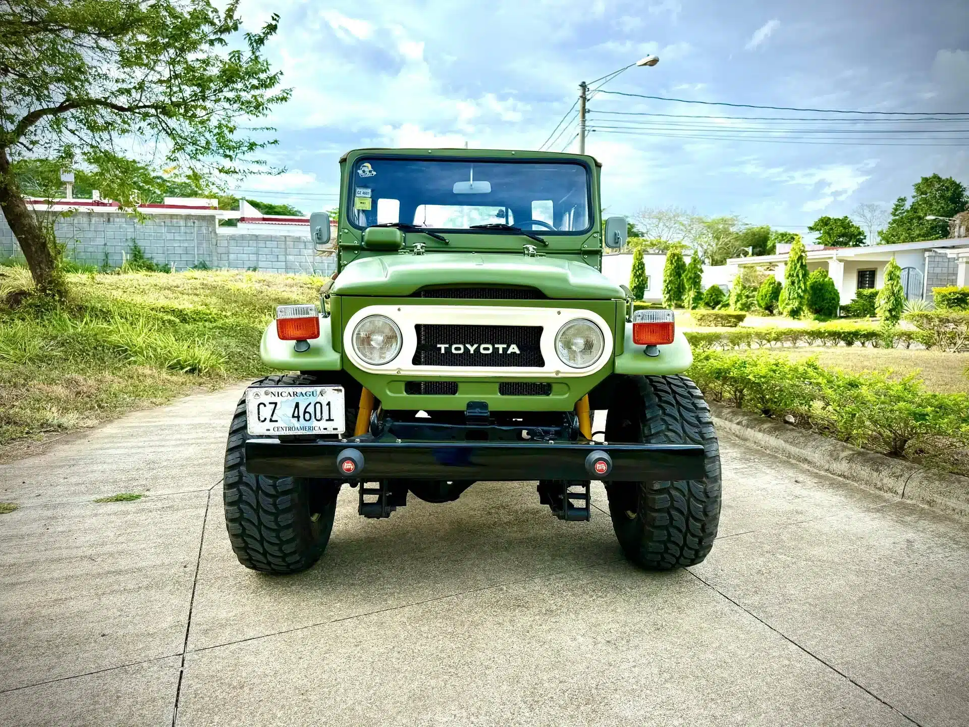 Vorderansicht eines grünen Toyota Land Cruiser von 1978 auf einer Straße, im Hintergrund Wohnhäuser und Bäume