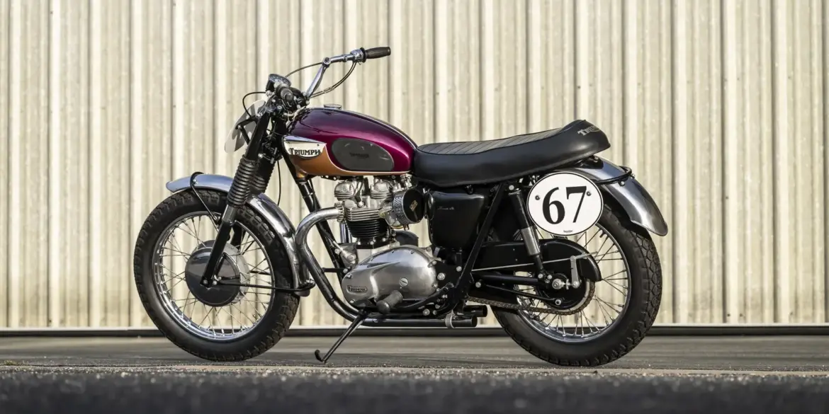 Restaurierte Triumph Bonneville TT Special von 1967 mit auberginefarbenem Tank, Edelstahl-Kotflügeln und Startnummer 67 vor einer Blechwand