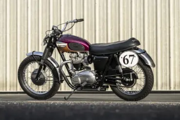 Restaurierte Triumph Bonneville TT Special von 1967 mit auberginefarbenem Tank, Edelstahl-Kotflügeln und Startnummer 67 vor einer Blechwand