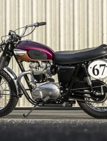 Restaurierte Triumph Bonneville TT Special von 1967 mit auberginefarbenem Tank, Edelstahl-Kotflügeln und Startnummer 67 vor einer Blechwand