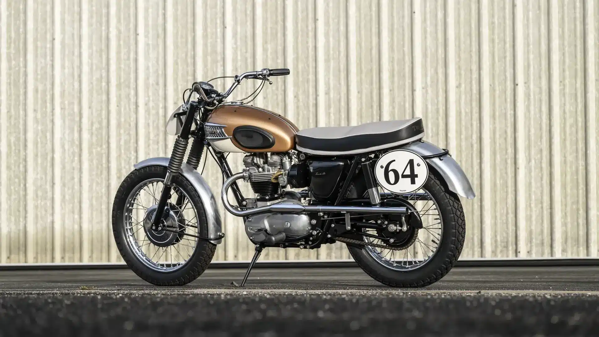 Restaurierte Triumph Bonneville TT Special 1964 im Seitenprofil mit Startnummer 64 vor einer hellen Wand.