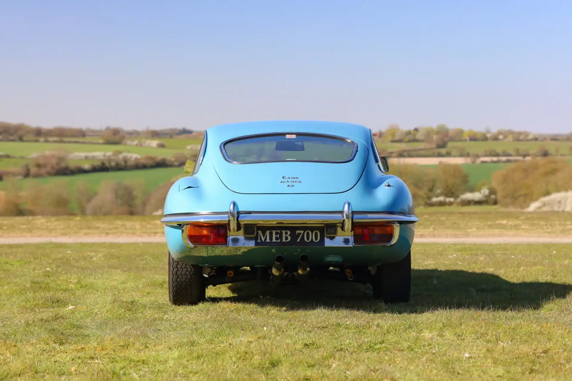Heckansicht eines hellblauen Jaguar E-Type 4.2 Series 2 Fixed Head Coupé, Baujahr 1969, mit dem Kennzeichen MEB 700 vor ländlicher Kulisse.