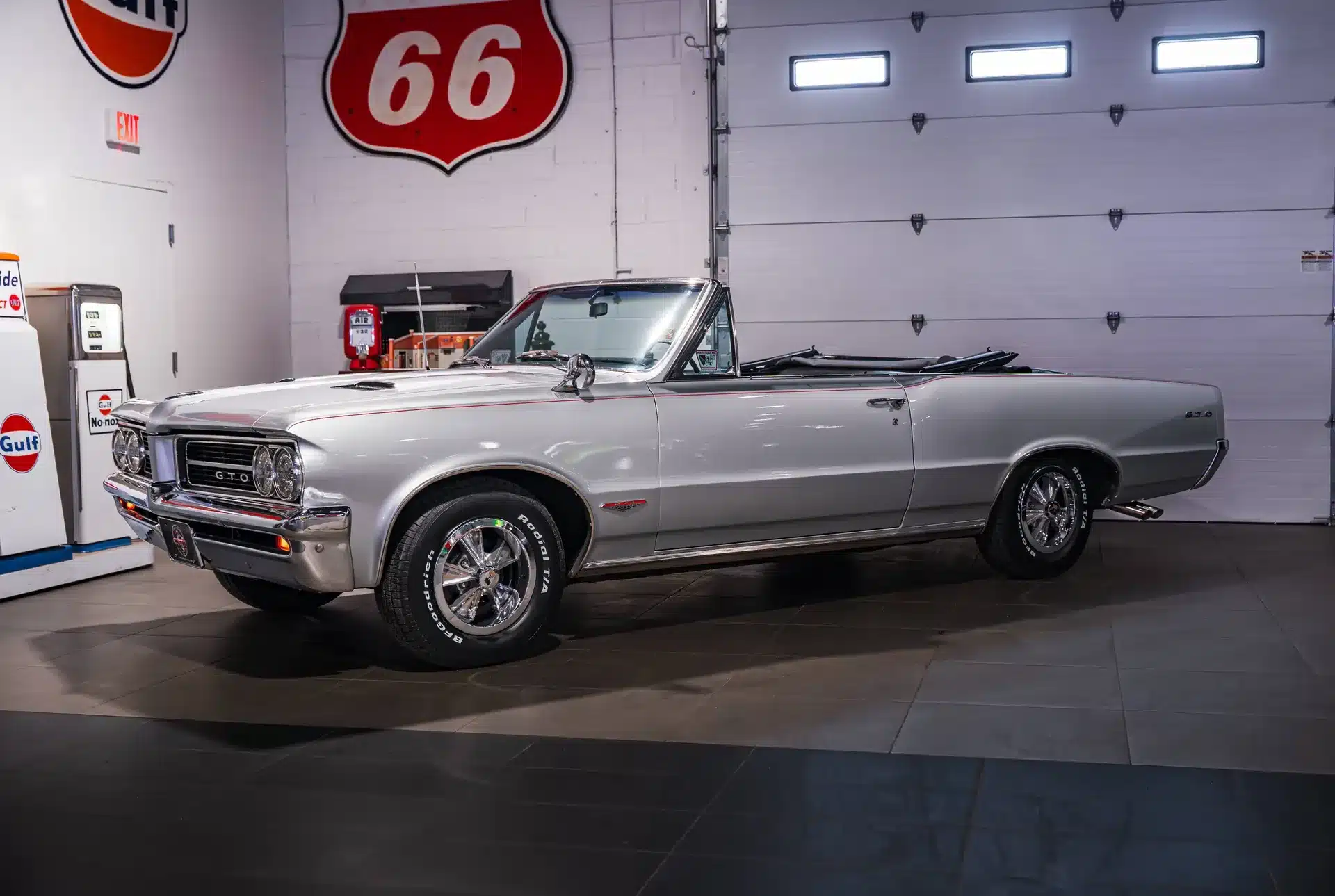 Silbernes Pontiac GTO Cabriolet Baujahr 1964 in einer Ausstellungshalle mit Retro-Tankstellen im Hintergrund.