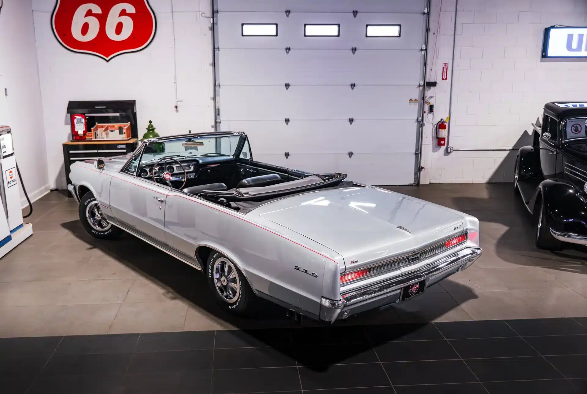 Ein silbernes 1964 Pontiac GTO Cabriolet mit schwarzem Interieur steht offen in einer klassischen Werkstatt mit Werkbank und historischen Zapfsäulen.