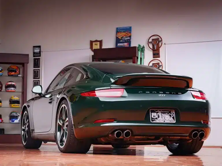 Porsche 911 Carrera S Club Coupé 2013 in Brewster Green von schräg hinten, mit Ducktail-Heckspoiler, in einer Garage mit Trophäen und Helmen.