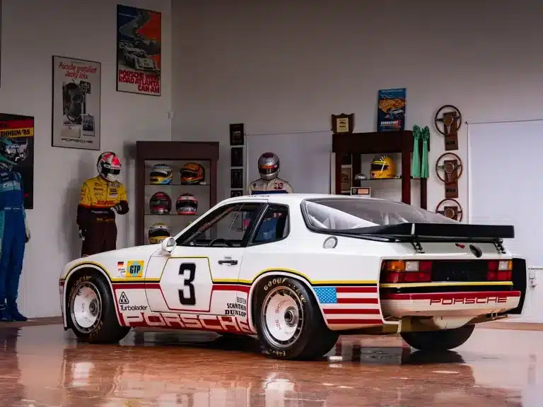 Porsche 924 GTP Rennwagen von 1980, weiß mit Startnummer 3, Seitenansicht, Ausstellung mit Motorsport-Postern und Helmen im Hintergrund.