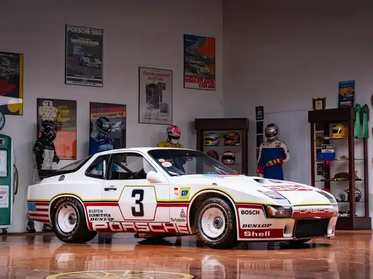 Porsche 924 GTP von 1980 mit Startnummer 3 in einer Sammlung, Seitenansicht vor Wand mit Rennpostern und Helmen.