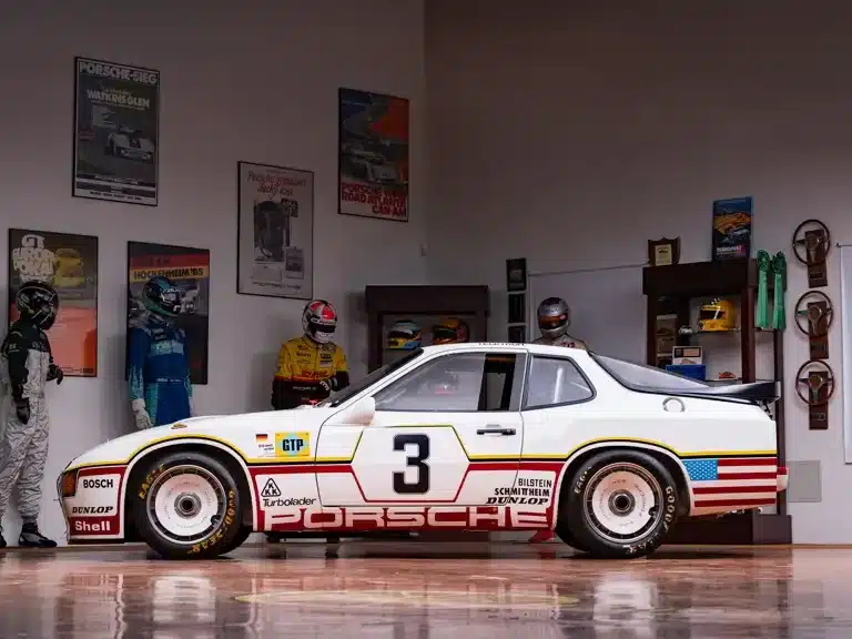 Seitenansicht des Porsche 924 GTP von 1980 mit Startnummer 3 im Ausstellungsraum, dekoriert mit Motorsport-Postern und Rennfahrer-Overalls.