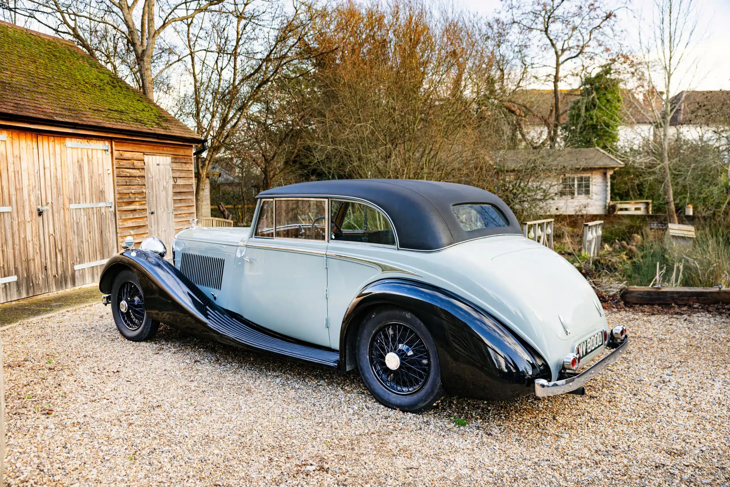 Bentley 4 1/4 Litre Coupé von 1936 in hellblau mit schwarzem Dach und Kotflügeln, auf einem Kiesweg vor einer Holzhütte geparkt.