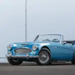 Austin-Healey 3000 Mk III BJ8 1965 in Blau, offen, vor heller Wand, schräg von vorn fotografiert.