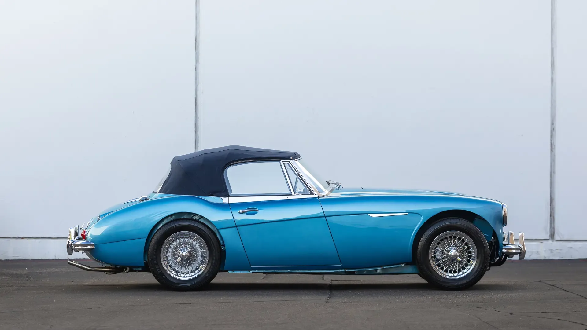 Blauer Austin-Healey 3000 Mk III BJ8 Cabriolet, Baujahr 1965, Seitenansicht mit Drahtspeichenrädern und geschlossenem Verdeck vor einer weißen Wand.