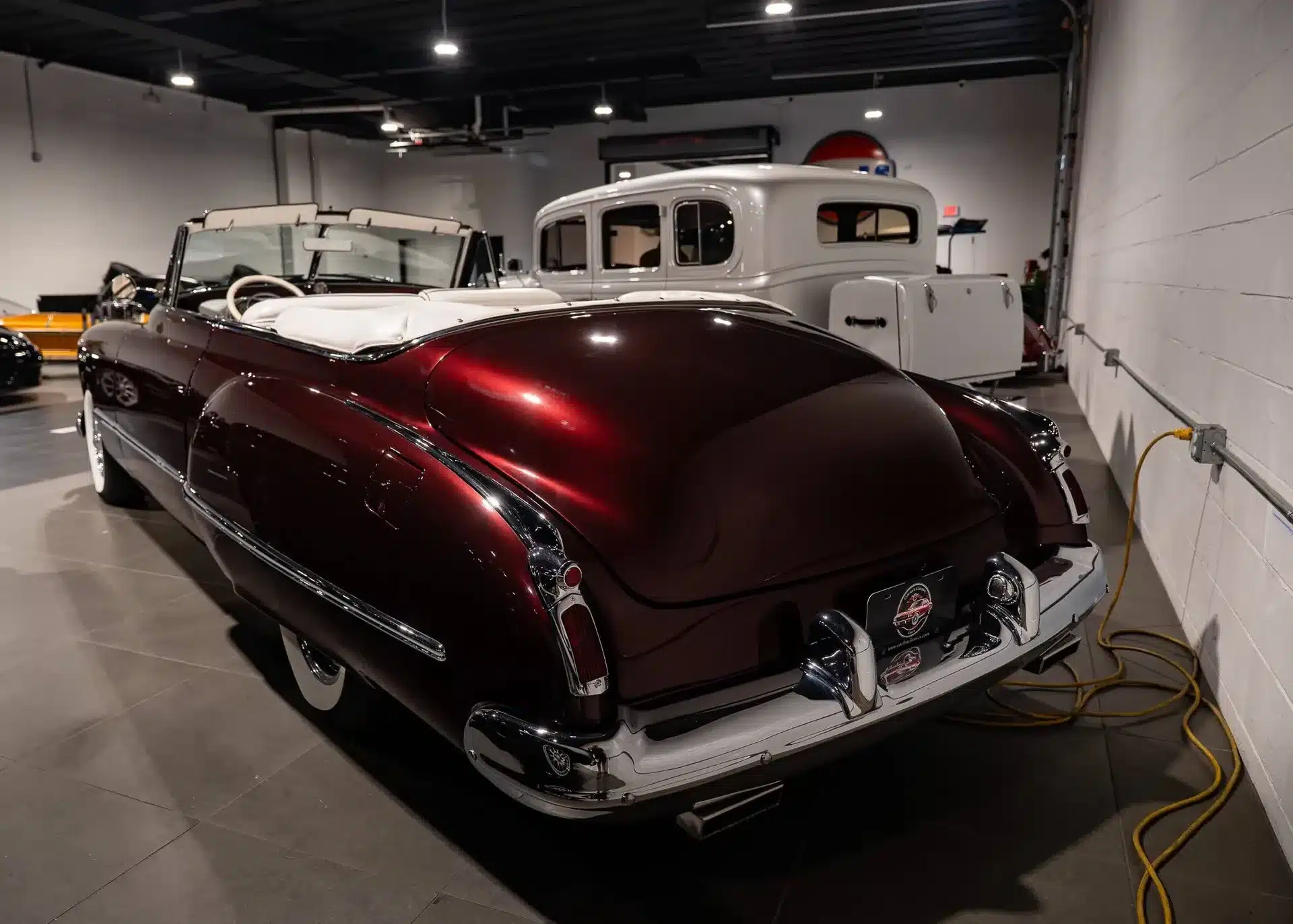 Burgunderrotes Oldsmobile 88 Cabriolet von 1948 mit weißem Interieur, Blick auf das Heck, umgeben von weiteren Oldtimern in einer Halle.