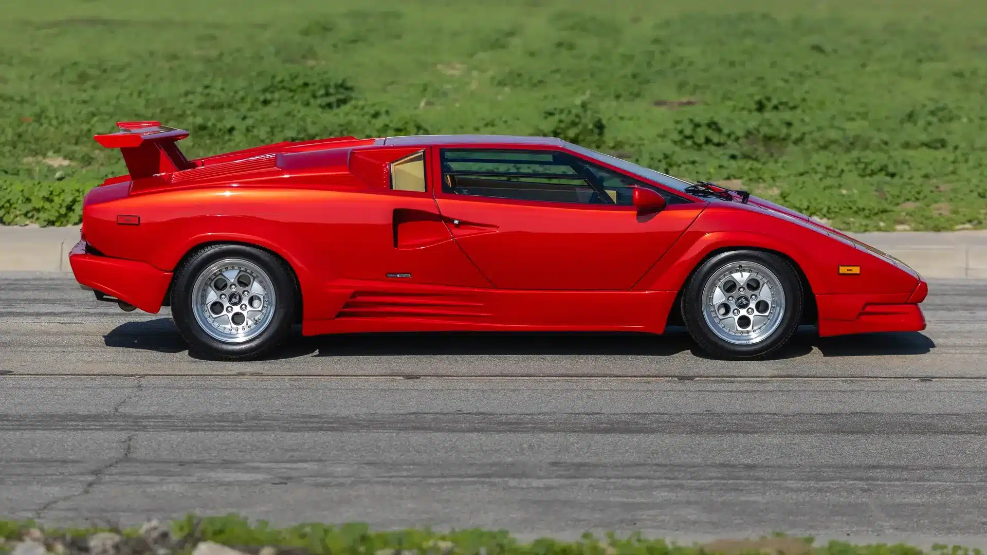 Seitenansicht eines roten Lamborghini Countach 25th Anniversary Edition von 1989 mit markantem Heckflügel und OZ-Racing-Felgen vor grünem Hintergrund.