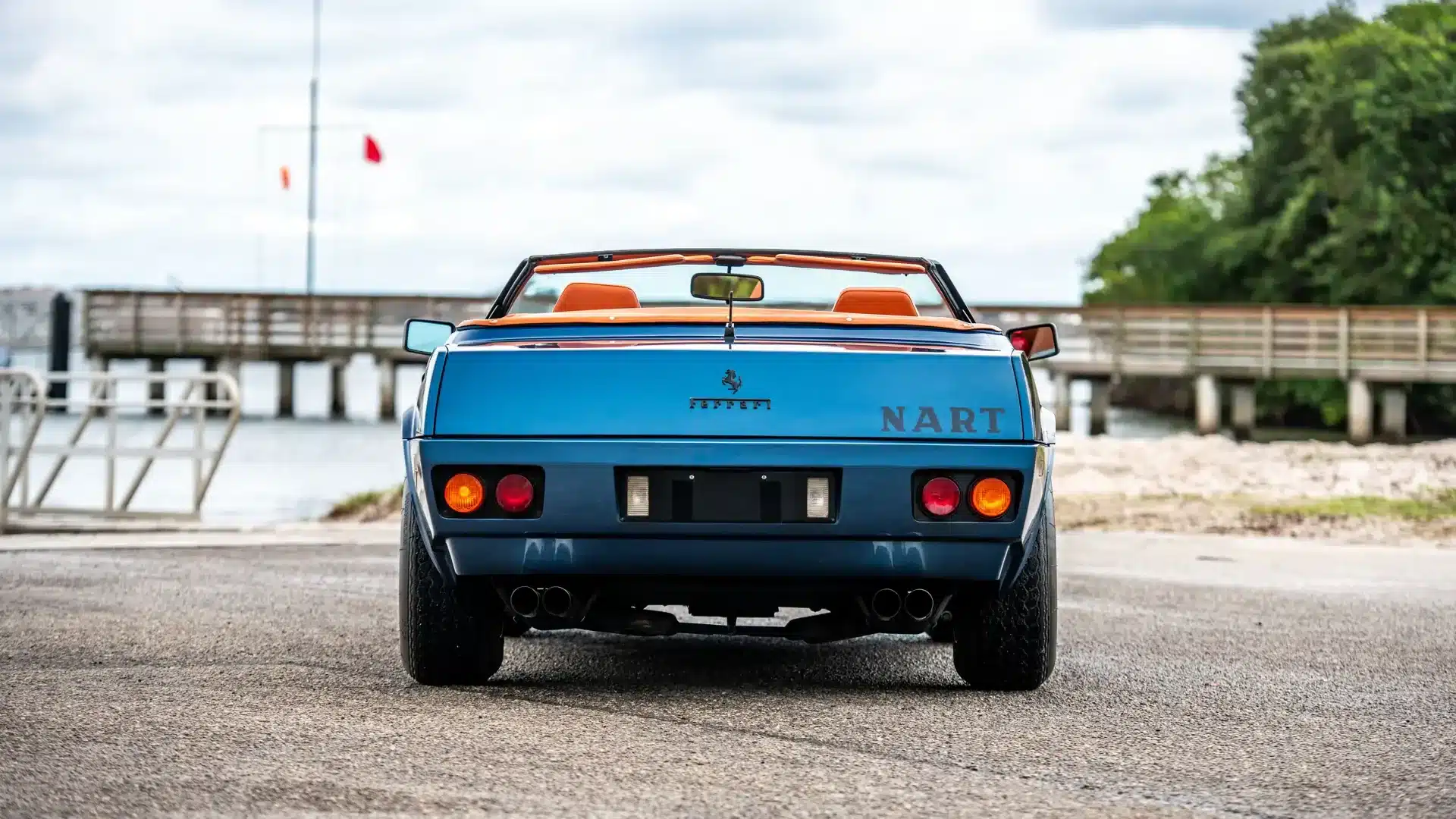 Rückansicht des blauen Ferrari 365 GTB/4 Daytona NART Spider mit geöffneter Verdeck und orangefarbenem Interieur, fotografiert an einer Uferpromenade.