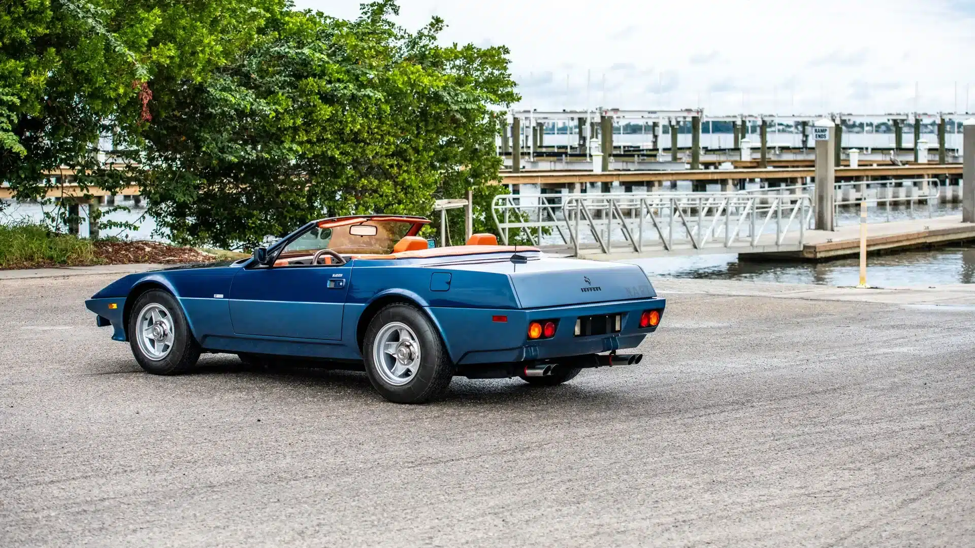 Rückansicht eines blauen Ferrari 365 GTB/4 Daytona NART Spider by Michelotti mit offenem Verdeck und orangefarbenem Interieur, am Hafen geparkt.