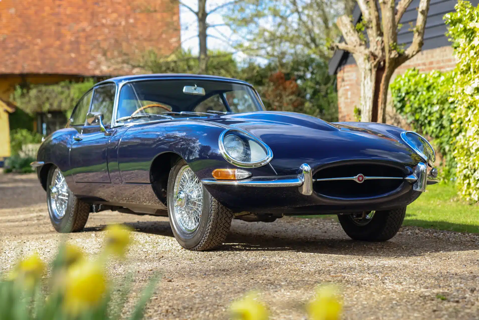 Seitenansicht eines Jaguar E-Type Series 1 4.2 Fixed Head Coupé von 1965 in Indigo Blue, geparkt auf einem Kiesweg im Grünen.