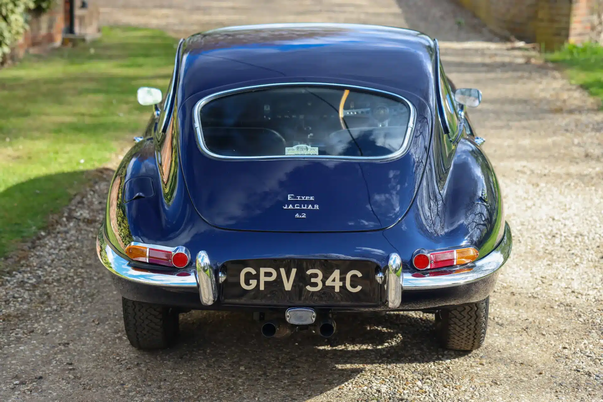Heckansicht eines blauen Jaguar E-Type Series 1 4.2 Fixed Head Coupé aus dem Jahr 1965 auf einer Schotterauffahrt.