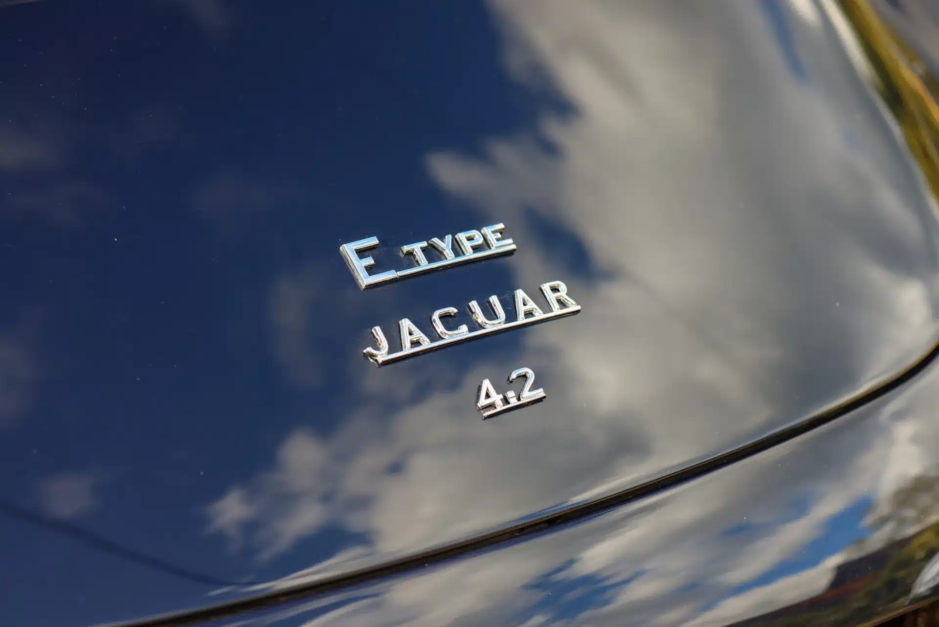 Nahaufnahme des Jaguar E-Type 4.2 Emblems auf der dunklen Motorhaube mit Wolkenspiegelung