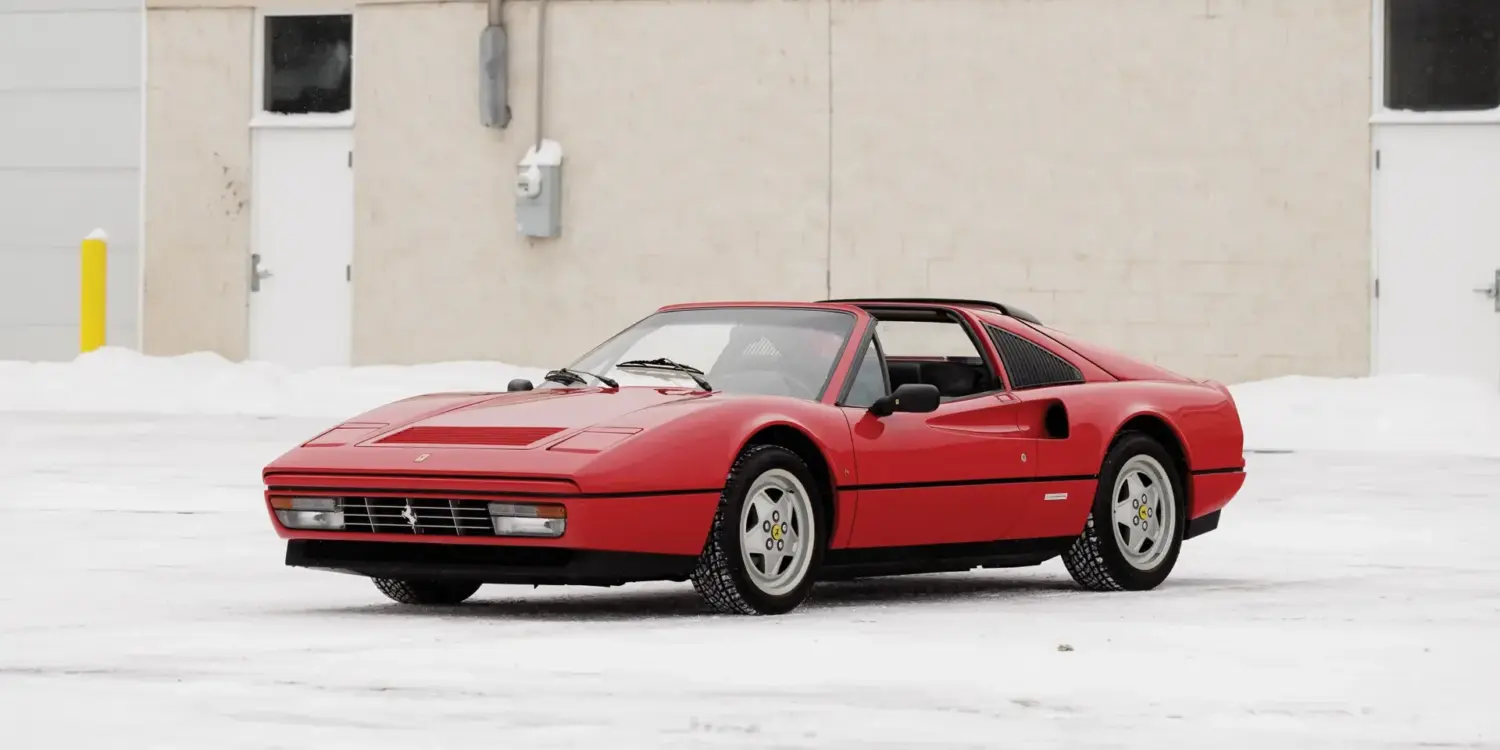 Ferrari 328 GTS von 1988 in Rosso Corsa, Targadach offen, geparkt auf schneebedecktem Boden vor beigem Gebäude