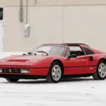 Ferrari 328 GTS von 1988 in Rosso Corsa, Targadach offen, geparkt auf schneebedecktem Boden vor beigem Gebäude