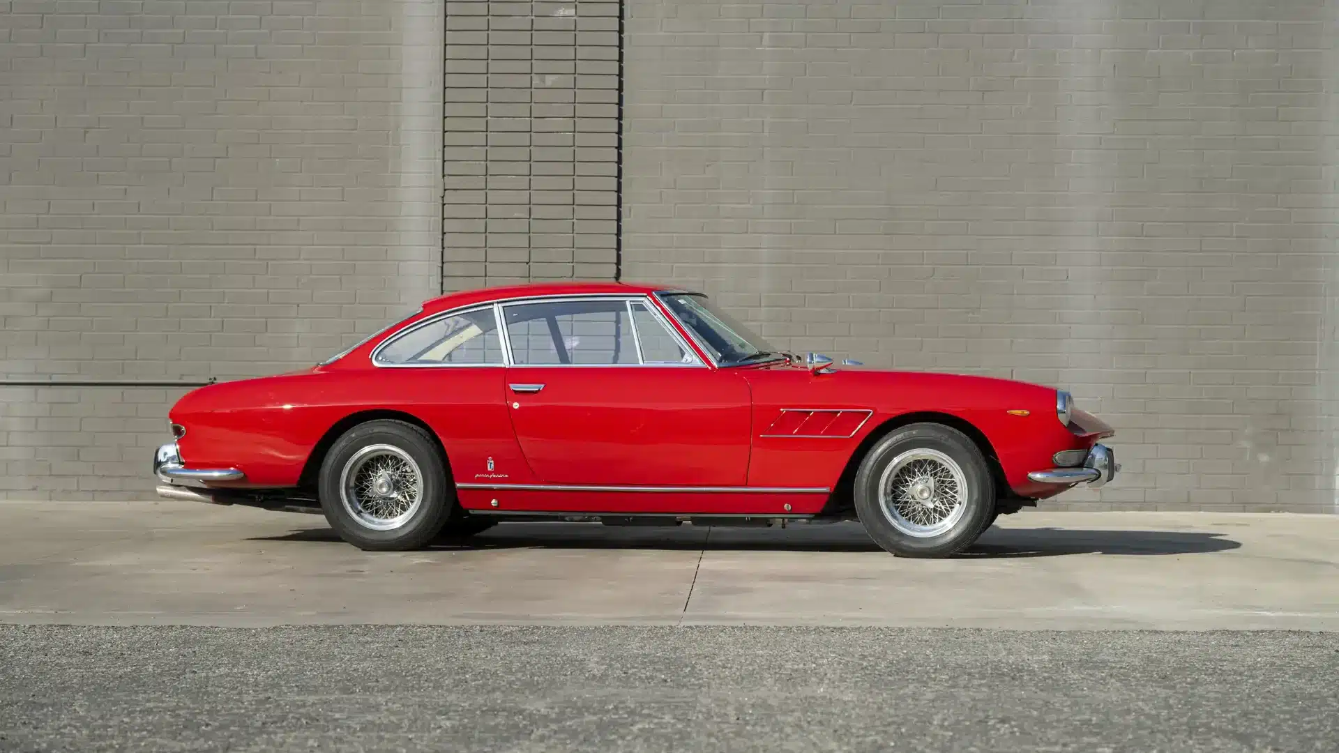 Seitenansicht eines roten Ferrari 330 GT 2+2 Coupé von 1966, parkend vor einer grauen Mauer.