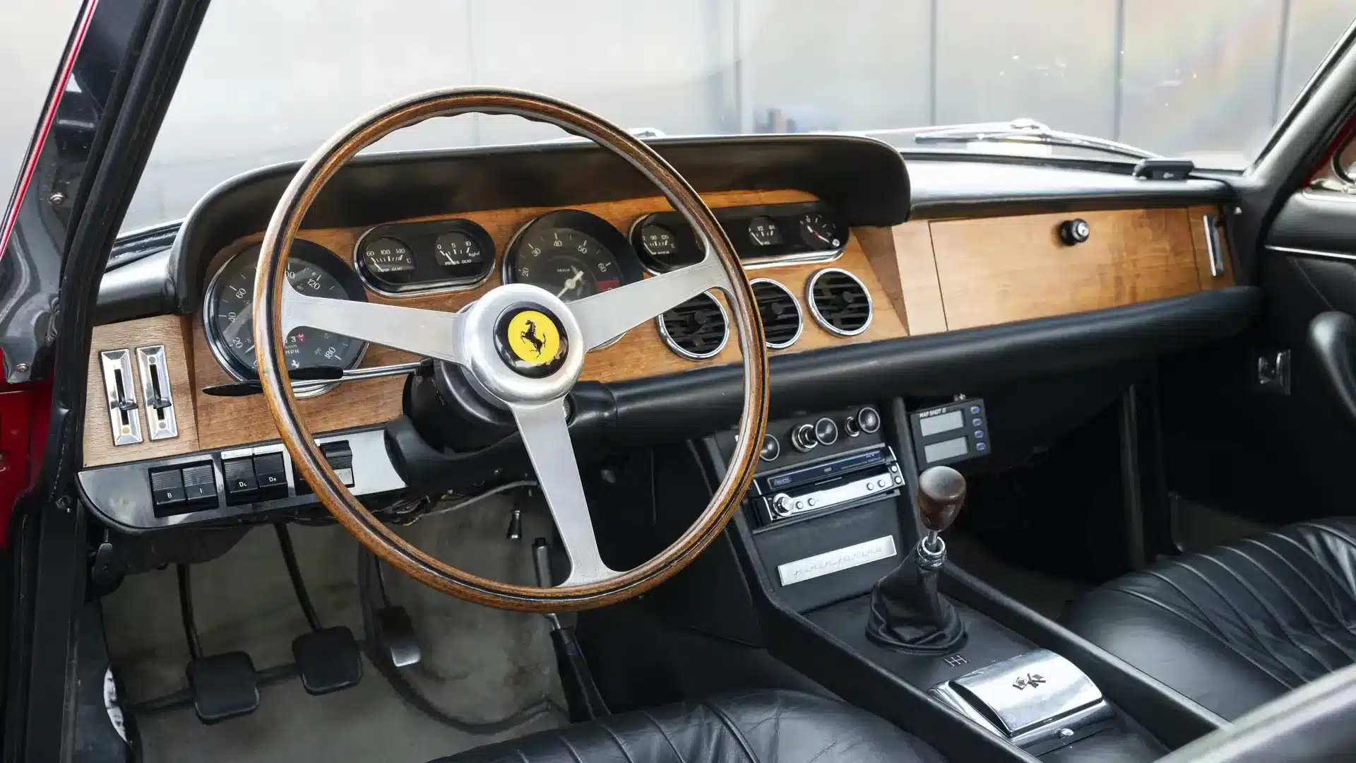 Lenkrad und Armaturenbrett eines Ferrari 330 GT 2+2 Series II, Baujahr 1966, mit Holzverkleidung und schwarzen Ledersitzen.