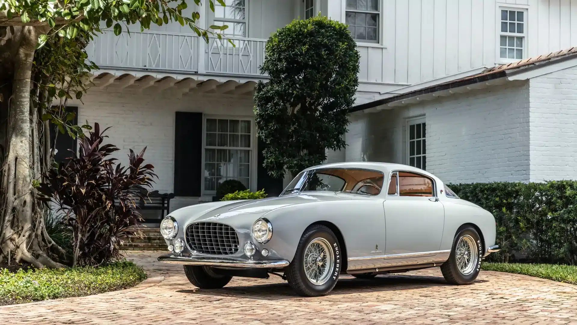 Ferrari 250 Europa GT Coupé von 1955 in Grau mit orangenem Interieur, geparkt vor einem weißen Gebäude mit Garten.