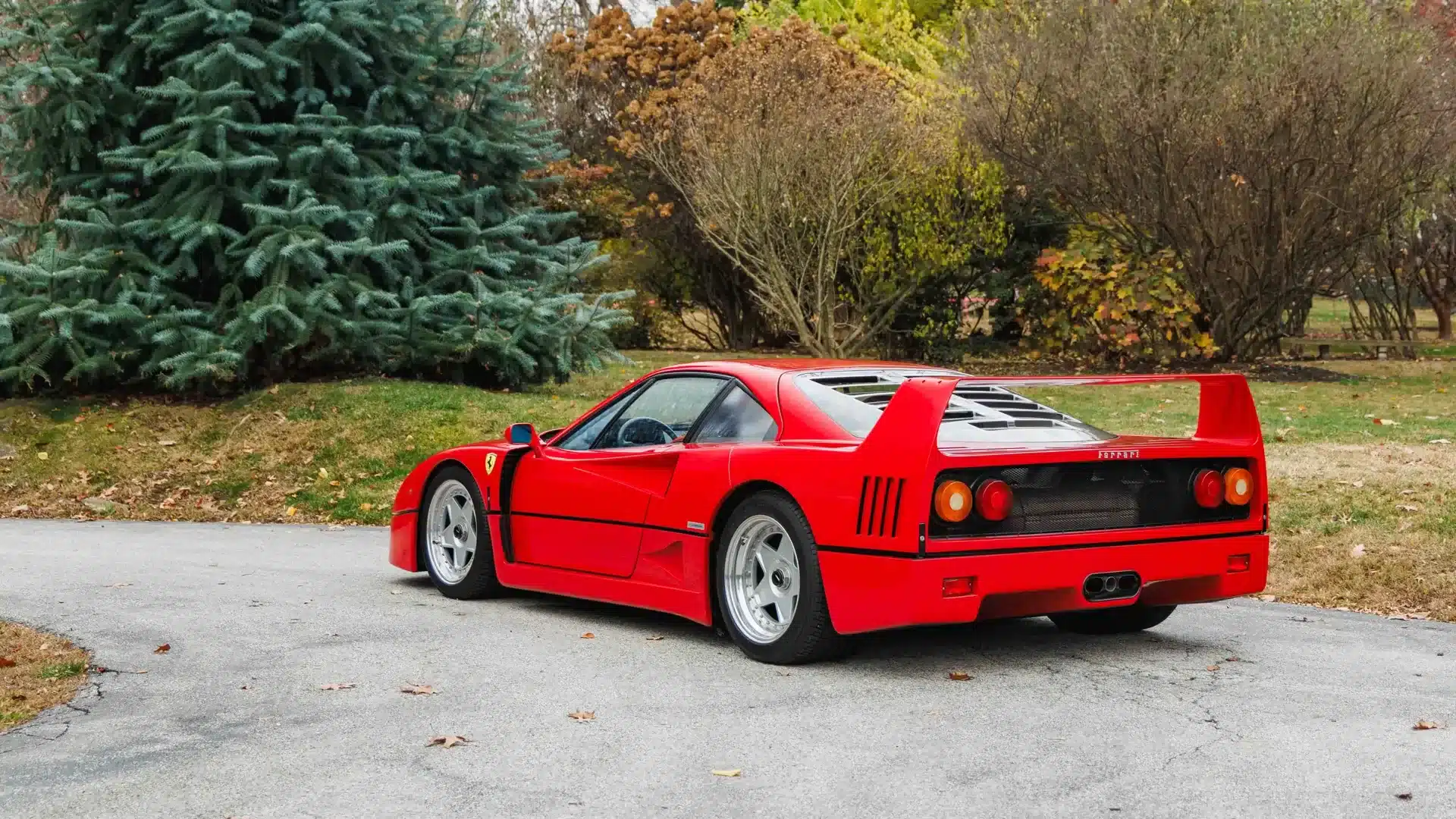 Roter Ferrari F40 aus dem Baujahr 1991 mit markantem Heckflügel, geparkt auf einer Einfahrt vor Bäumen.