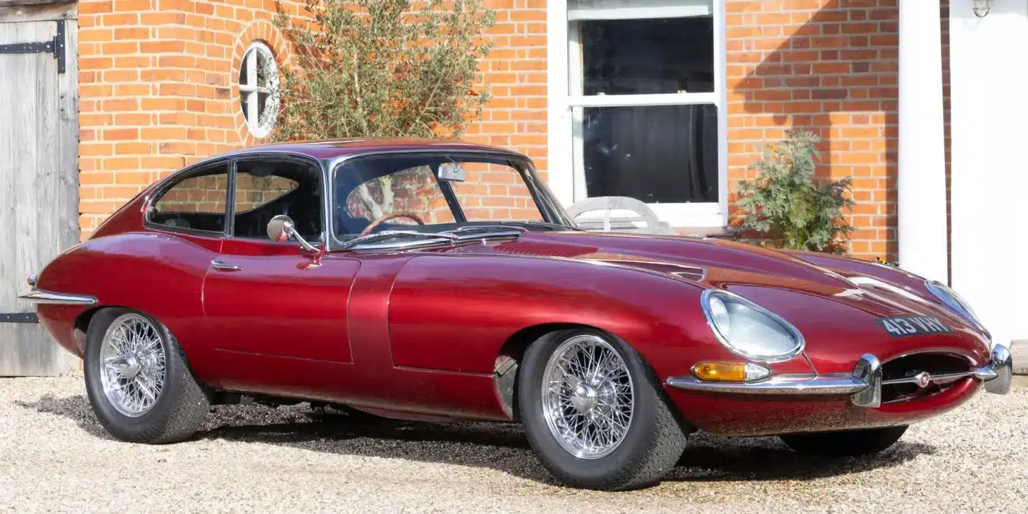 Jaguar E-Type 3.8 Litre Coupé von 1962 in Burgunderrot steht vor einem historischen Landhaus.