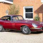 Jaguar E-Type 3.8 Litre Coupé von 1962 in Burgunderrot steht vor einem historischen Landhaus.
