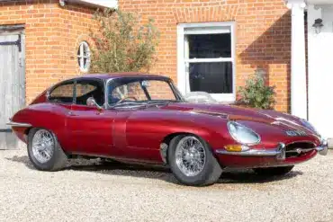 Jaguar E-Type 3.8 Litre Coupé von 1962 in Burgunderrot steht vor einem historischen Landhaus.