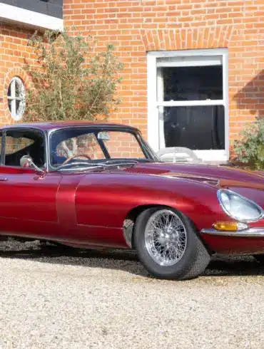 Jaguar E-Type 3.8 Litre Coupé von 1962 in Burgunderrot steht vor einem historischen Landhaus.