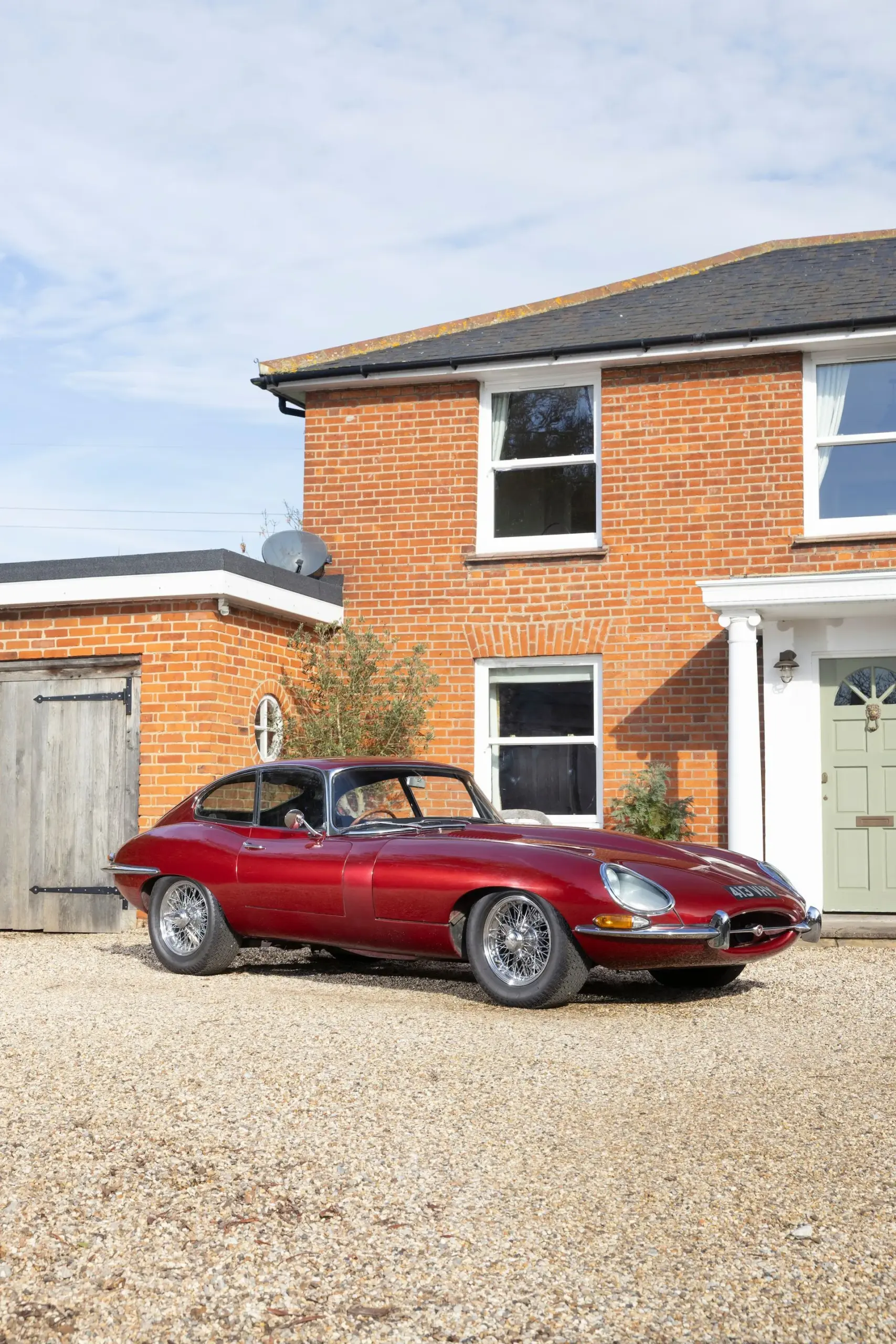 Jaguar E-Type 3.8 Litre Coupé, Baujahr 1962, in Burgunderrot vor einem Ziegelhaus geparkt.