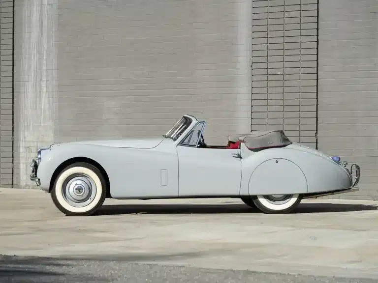 Jaguar XK 120 Drophead Coupé von 1953 in Birch Grey, Stoffverdeck geschlossen, Seitenansicht vor einer grauen Mauer